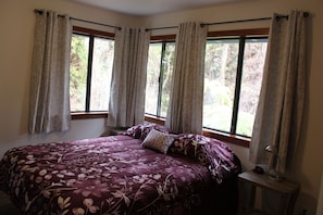 A bedroom with a large bed, two windows with curtains, and a bedside table with a lamp.