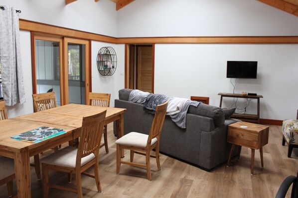 A living room with a wooden dining table, chairs, a grey sofa, and a TV on a wooden stand.