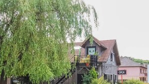 Una casa de madera rústica con una escalera que conduce a un balcón, rodeada de abundante vegetación y un letrero que dice 'OPEN'.