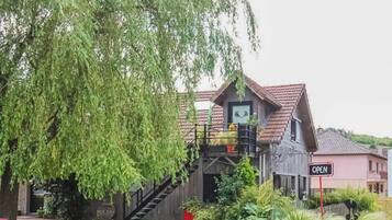 Tunnelmallinen, maalaishenkinen puutalo, jossa on portaikko, joka johtaa parvekkeelle. Talon ympärillä on vehreää kasvillisuutta, ja sen edessä on kyltti, jossa lukee: ”AVOIN”.