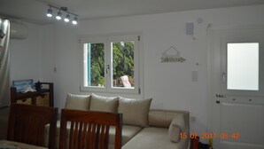 A room with a sofa, a dining table with chairs, a window with a view of greenery, and a door with a light switch.