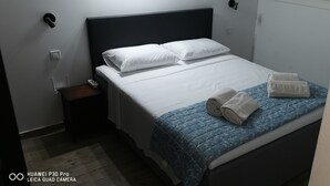 A neatly made bed with white linens and a blue floral duvet, a small bedside table with a phone and a lamp, and a wall-mounted light fixture.
