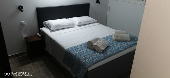 Uma cama bem arrumada com lençóis brancos e um edredom floral azul, uma pequena mesa de cabeceira com um telefone e uma luminária, e um aplique na parede.