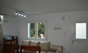 Ein Zimmer mit einer Couch, einem Esstisch mit Stühlen, einem Fenster mit Blick ins Grüne und einer Tür mit Lichtschalter.