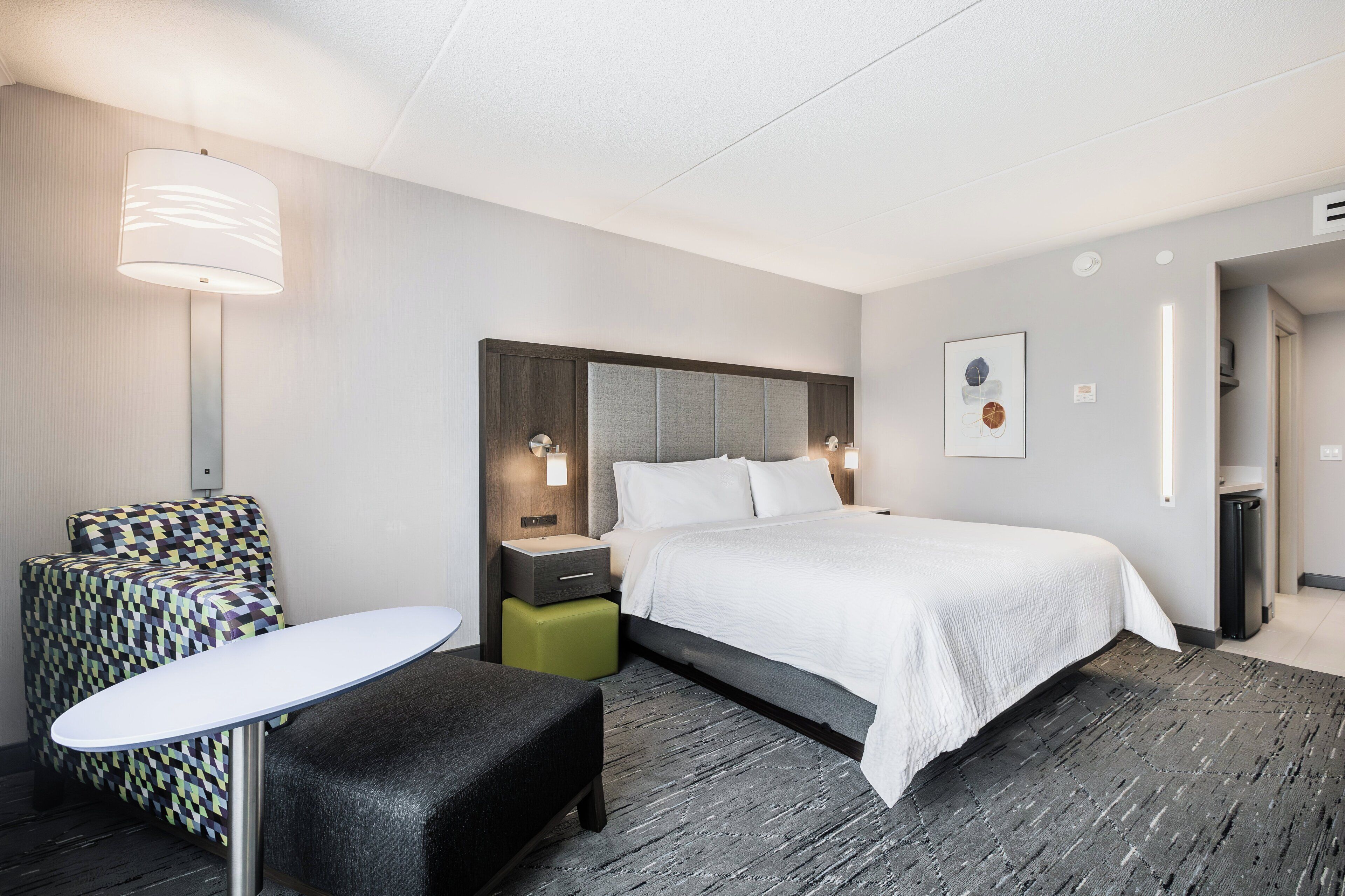 Quarto de hotel moderno com uma cama grande, uma mesinha, uma poltrona estampada e uma mesa de cabeceira com abajur.