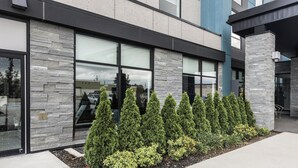 A modern building with large windows, a stone facade, and a landscaped area with coniferous trees and shrubs. - Holiday Inn Express & Suites Toronto Airport South by IHG (Toronto)