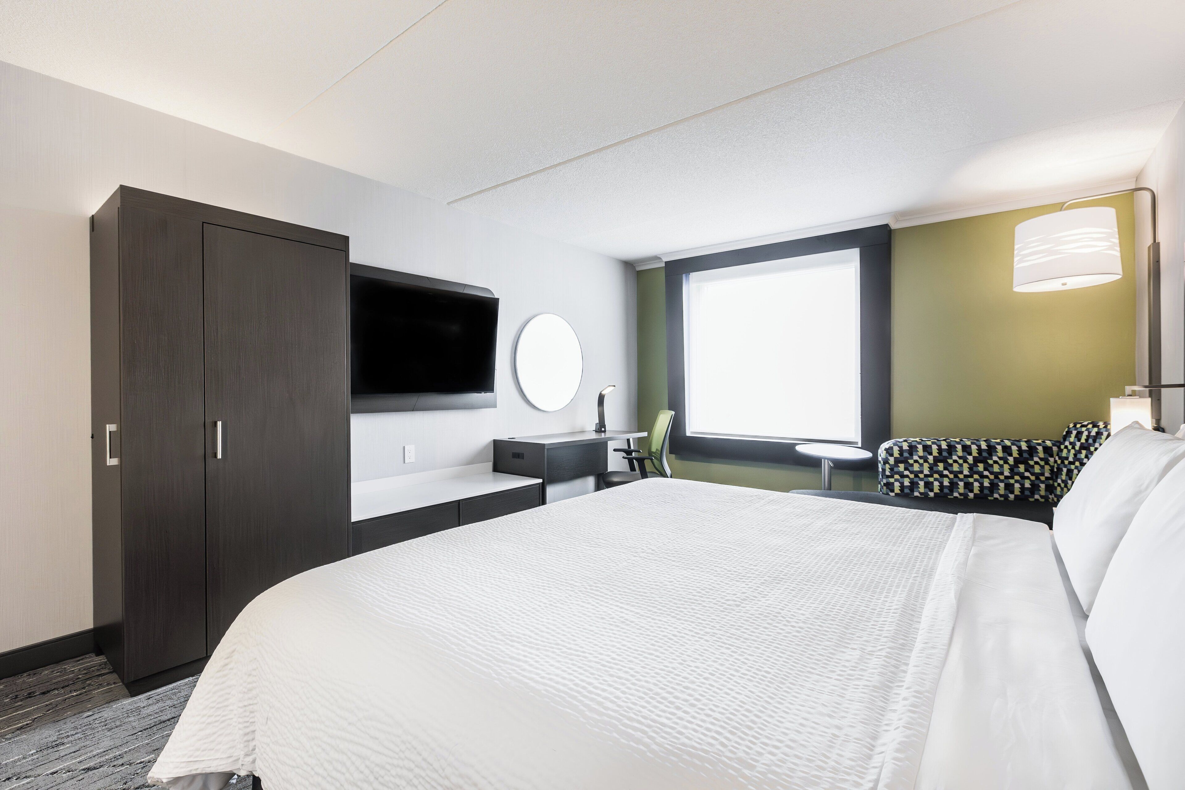 Ein Hotelzimmer mit einem großen Bett, einem Schreibtisch, einem Flachbildfernseher, einem runden Spiegel und einem Fenster mit Blick.
