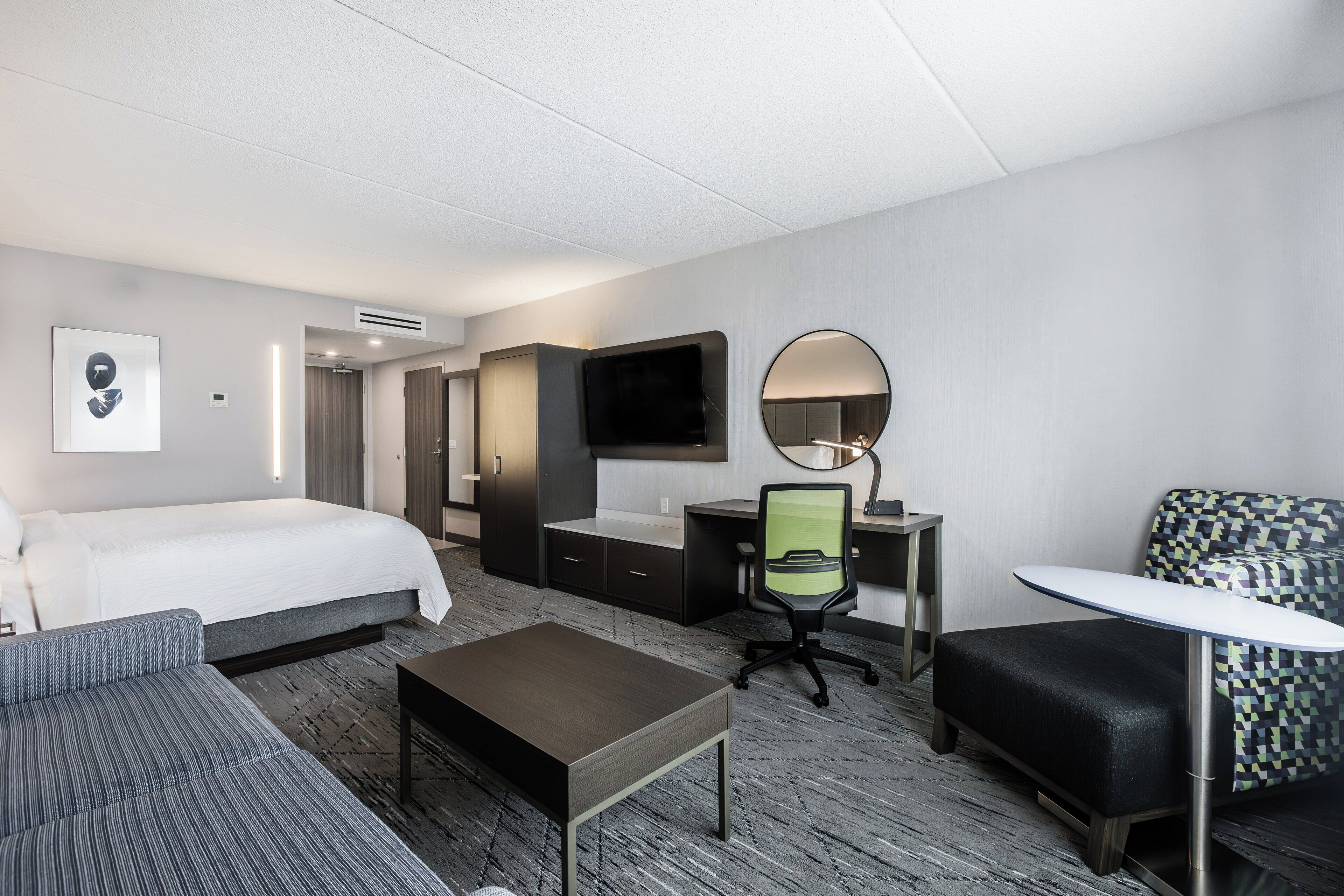 A hotel room with a bed, a desk, a chair, a small table, a mirror, and a TV.
