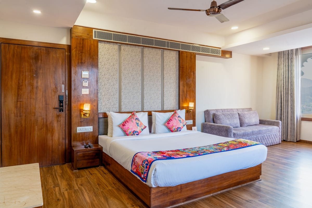 A modern hotel room with a wooden bed, a grey sofa, and a large window with curtains.