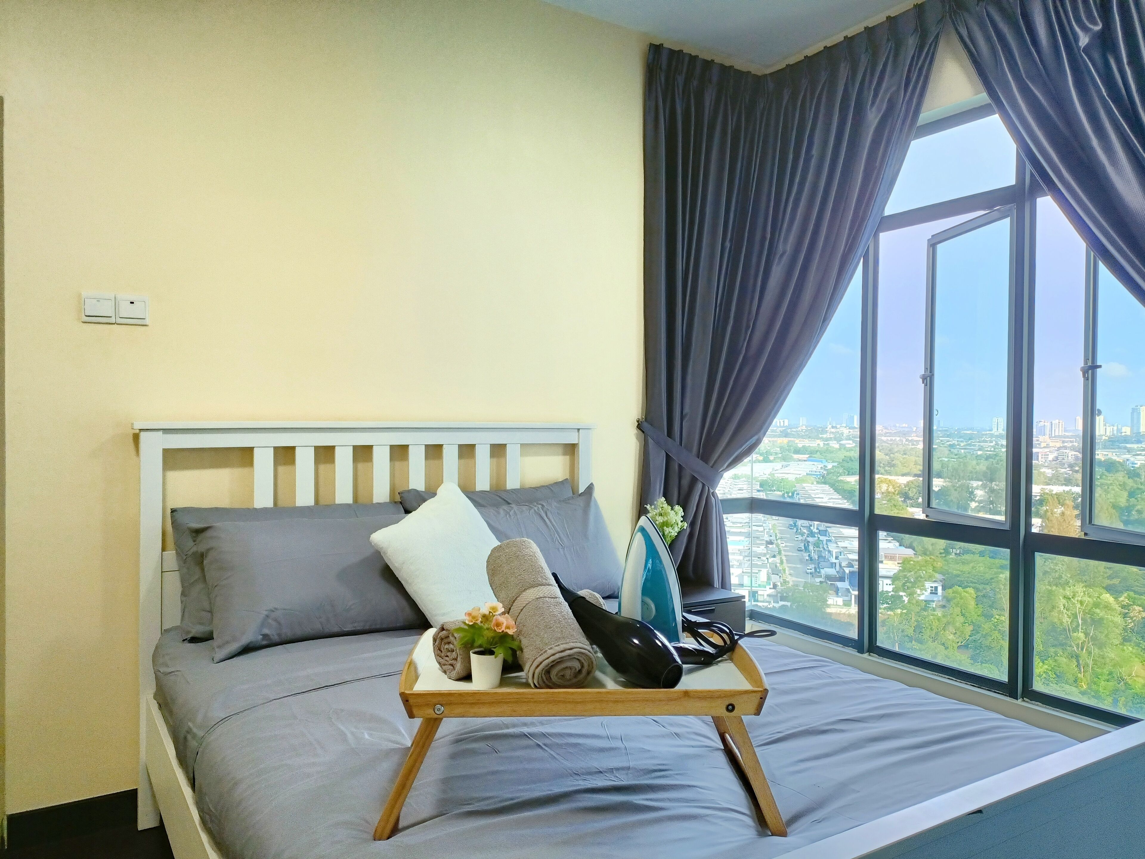 A bed with grey bedding, a wooden tray with towels and a small plant, and a city view through a large window with dark curtains.