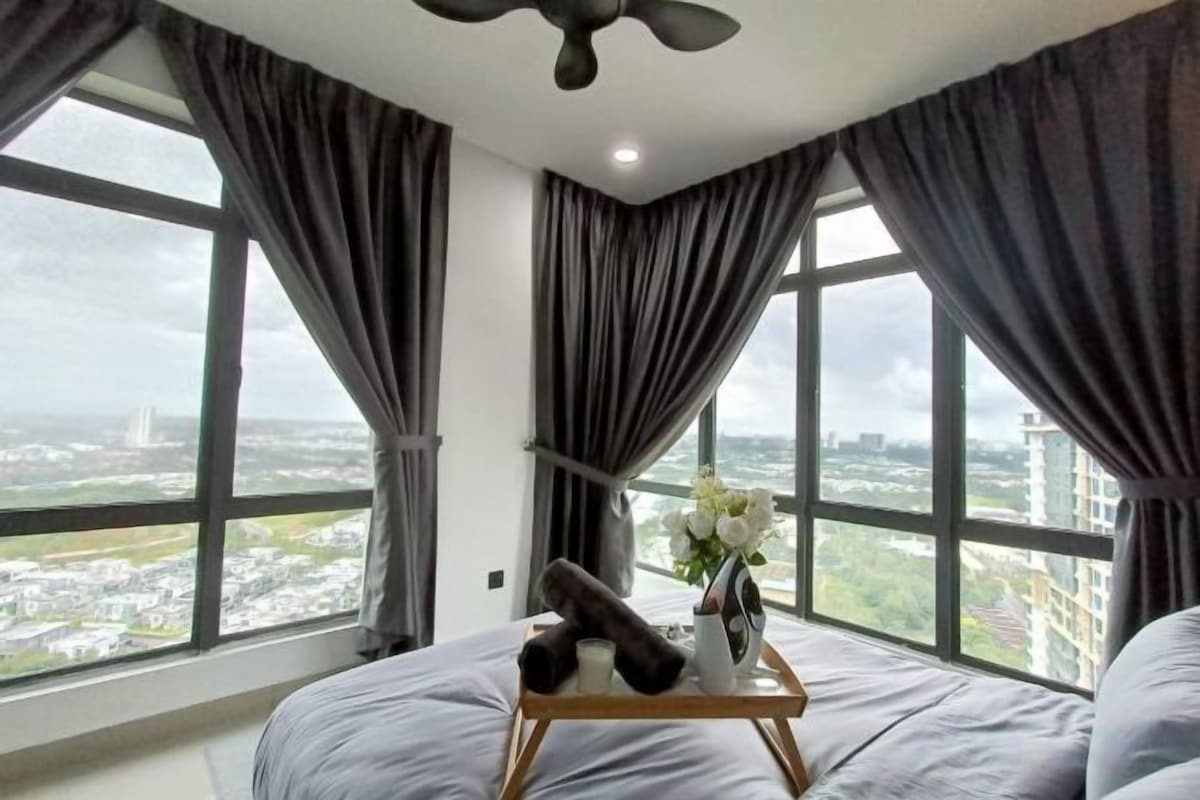 Quarto com cama, mesa de cabeceira com um vaso e controle remoto, e vista para a cidade através de janelas amplas.