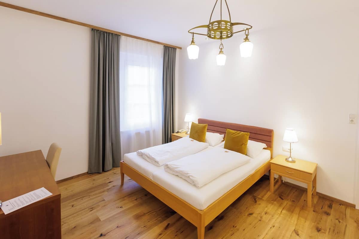 Habitación de hotel con una cama de madera, dos mesitas de noche con lámparas, un escritorio con una silla y un ventanal con cortinas.