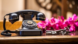 Un vieux téléphone à cadran rotatif, posé sur une surface en bois, avec des touches et des fleurs roses en arrière-plan.