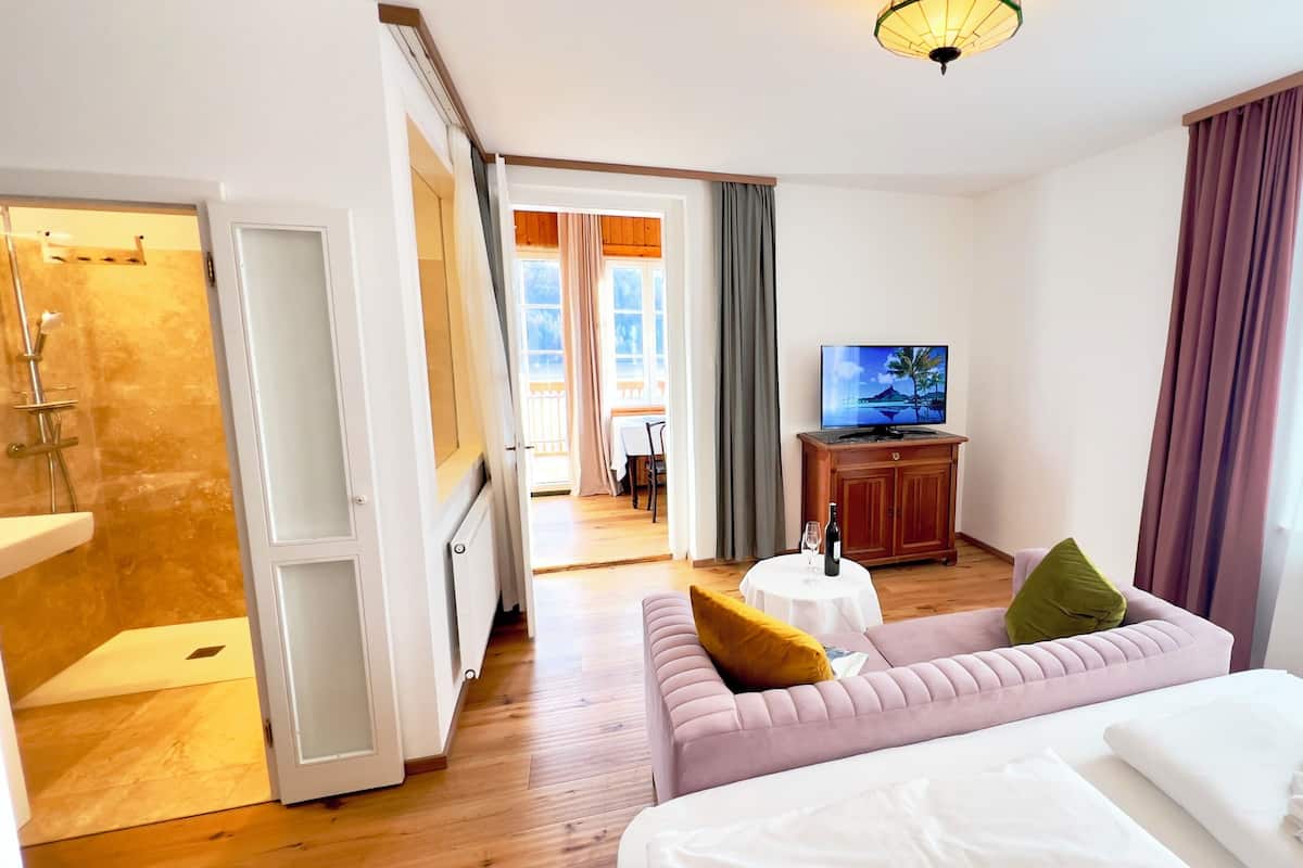 Habitación de hotel con cama, sofá, televisor, una mesita con una botella y vaso, y baño con ducha.