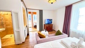 A hotel room with a bed, a sofa, a television, a small table with a bottle and glass, and a bathroom with a shower. - Hotel am See - Seeresidenz (Altaussee)