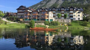 Une rangée d’immeubles à étages, dotés de balcons, s’étend le long d’un lac paisible dont les eaux reflètent le paysage, avec une montagne en arrière-plan.