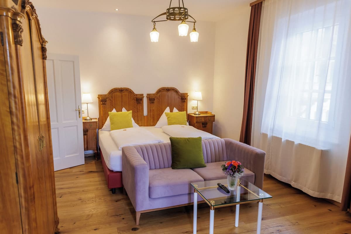 Una habitación de hotel con una cama, dos mesitas de noche con lámparas, un sofá, una mesa de centro de vidrio con un jarrón y un armario de madera.
