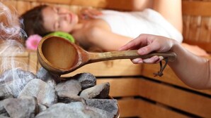 Un sauna avec des bancs en bois et une personne tenant une louche en bois au-dessus d’un tas de pierres.