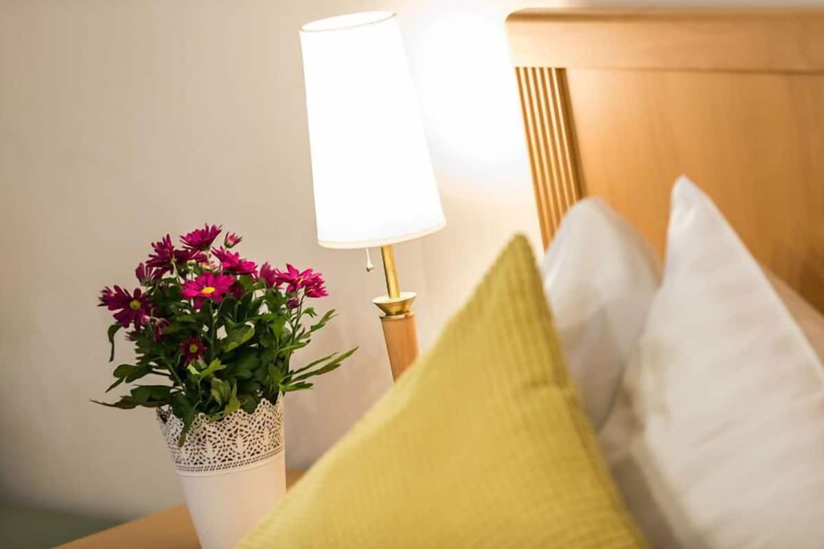 Una cama con almohadas blancas y amarillas, un florero blanco con flores rosas y una lámpara de noche con pantalla blanca.