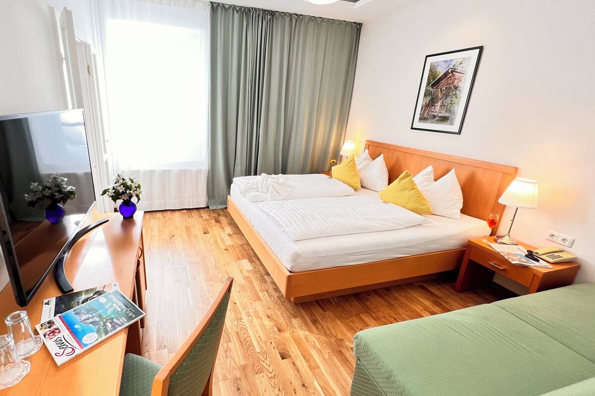 Habitación de hotel con cama, mesita de noche, lámpara, televisor y ventana con cortinas.