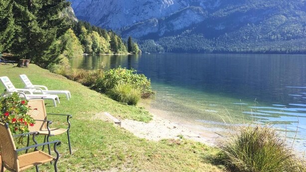 Une vue sur le lac, avec des montagnes en arrière-plan, une zone herbeuse avec des chaises longues et un lac aux eaux limpides.