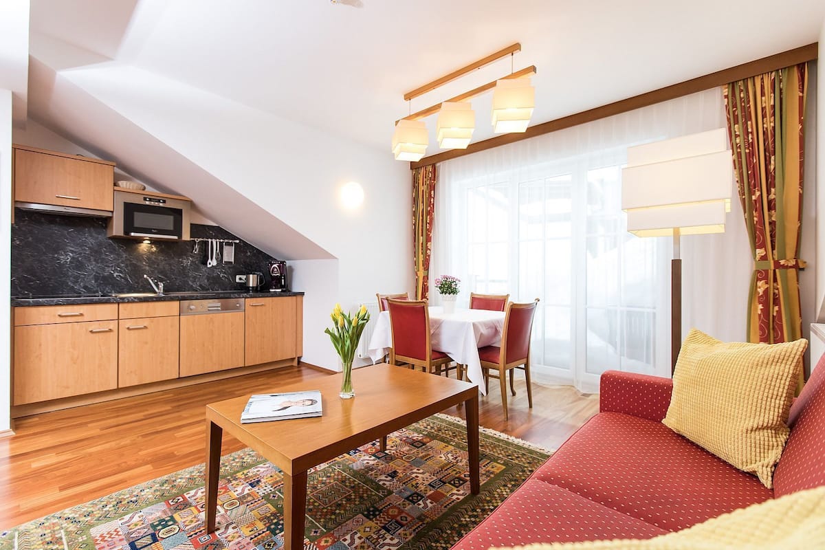 Una sala moderna con sofá, mesa de centro, mesa de comedor con sillas, zona de cocina con microondas y un amplio ventanal con cortinas.