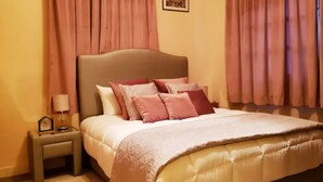 Cama bem arrumada com cabeceira bege e várias almofadas, mesa de cabeceira com abajur e quadro pendurado na mesinha de noite.