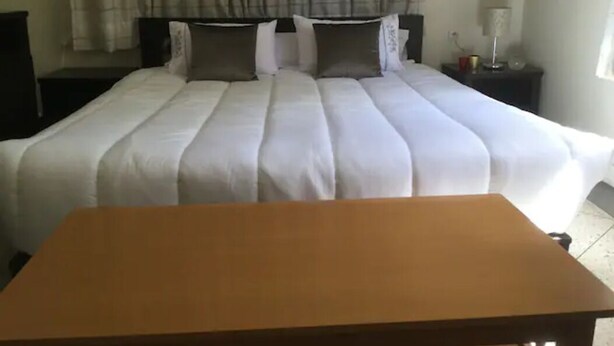 Uma cama bem arrumada com um edredom branco e várias almofadas, uma mesa de cabeceira de madeira com um abajur e uma janela com cortinas.
