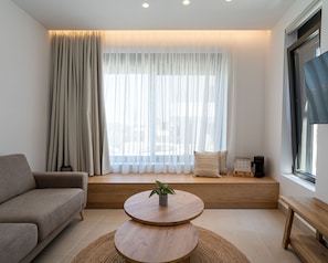 A modern living room with a sofa, a round wooden coffee table with a plant, a bench, and a large window with sheer curtains. - KoNoSo Luxury Apartments (Heraklion)