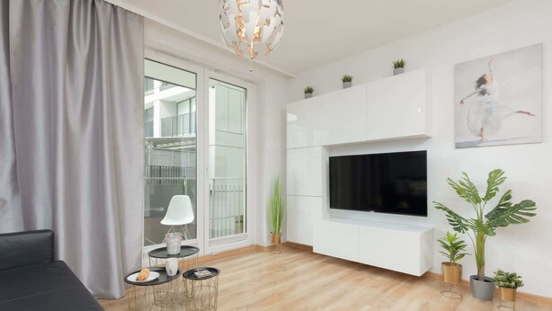 A modern living room with a black sofa, a small round table with a lamp, a TV mounted on the wall, and potted plants.