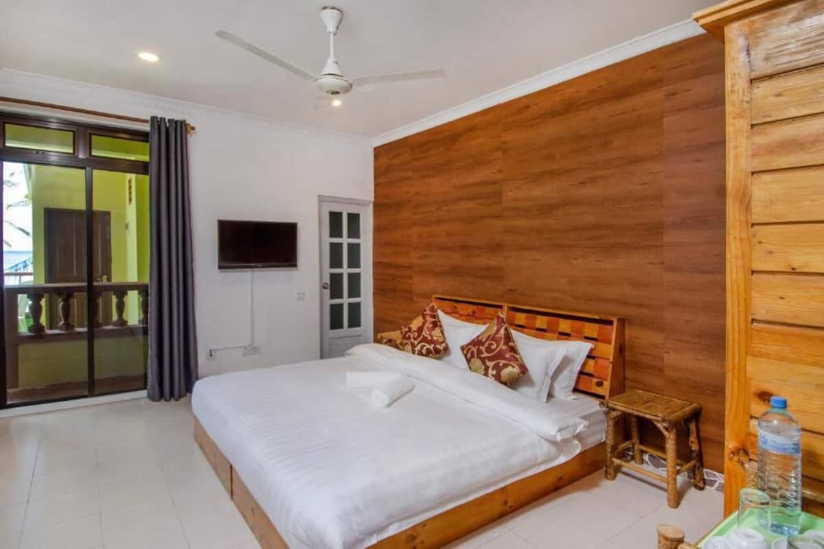A bedroom with a wooden headboard, a bed with white linens, a small wooden stool, a television on the wall, and a sliding glass door leading to an outdoor area.