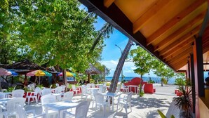 Un café frente a la playa con sillas y mesas de plástico blanco, sombrillas y vistas al océano.