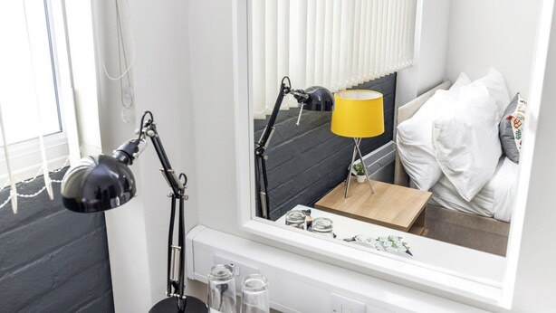 A hotel room with a bed, a bedside table with a lamp, a mirror, and a window with blinds.