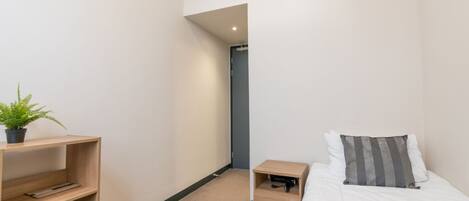 A bedroom with a bed, bedside table, and a shelf with a plant and cups.