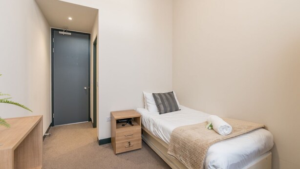 A small, clean bedroom with a single bed, a bedside table, a nightstand, and a door. - JOIVY Meadows Park Ensuite Single Room (Edinburgh)