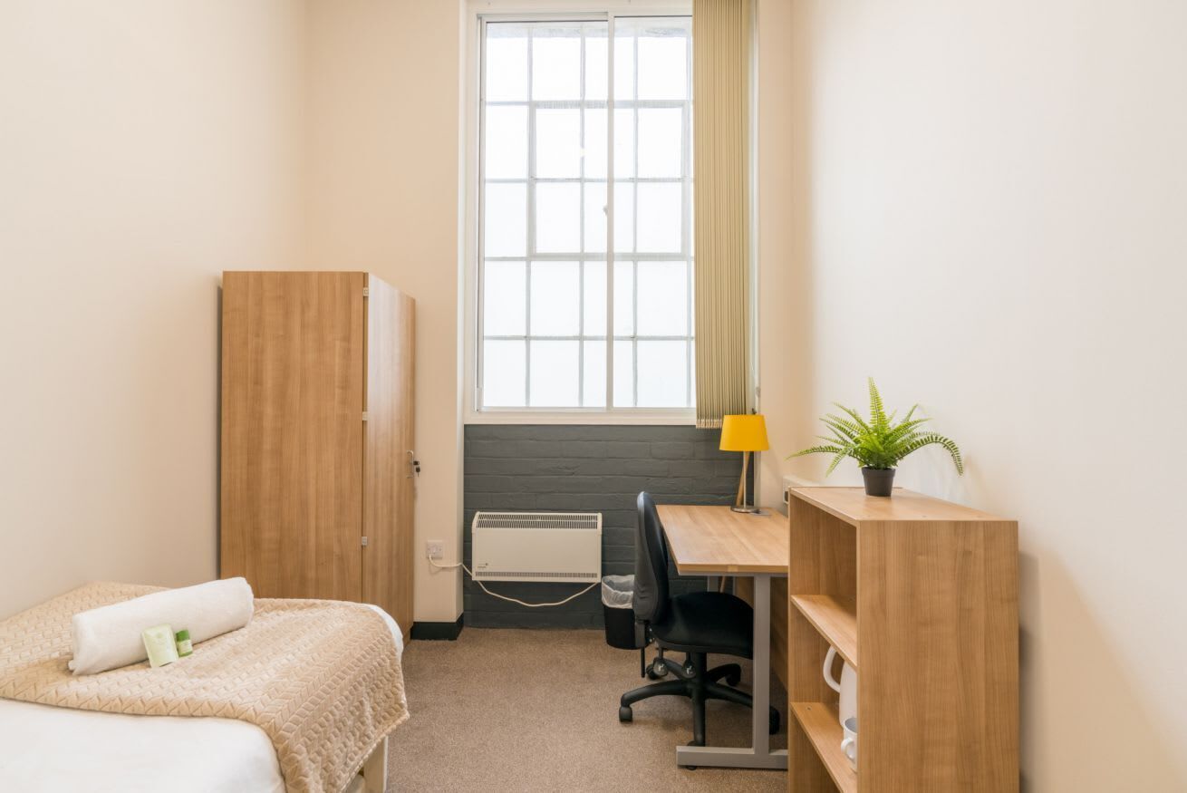 A small, well-lit room with a bed, a desk, a chair, a wardrobe, and a window with curtains.