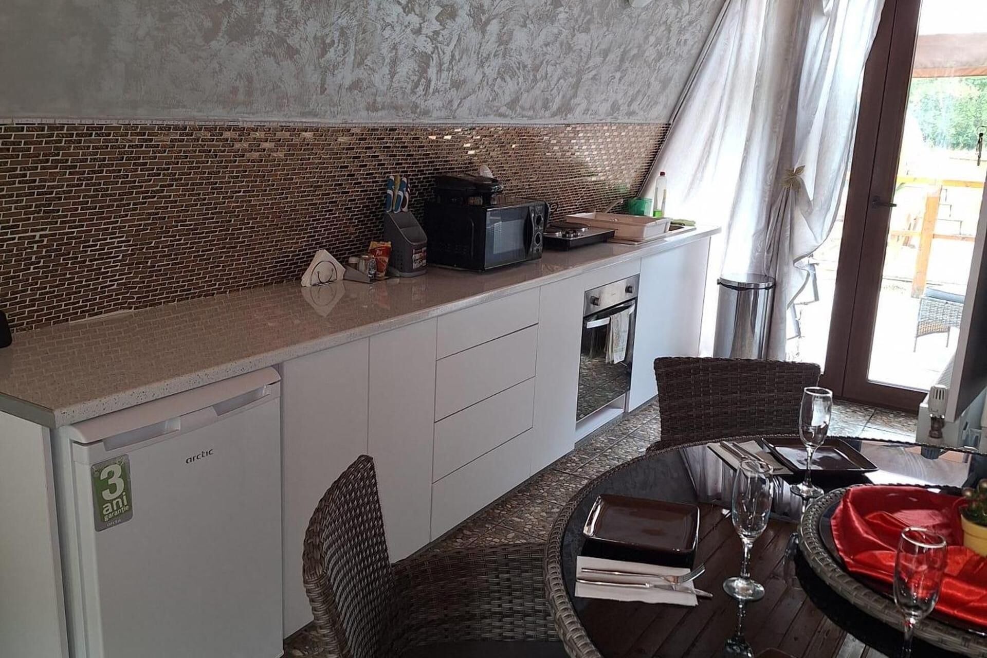 A compact kitchen with a brick backsplash, a microwave, and a small oven. There is a dining area with wicker chairs and a table set for two, featuring wine glasses and a red napkin.