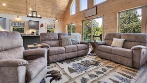 A spacious living room with two sofas, a coffee table, and a rug with a forest design. There are large windows providing natural light and a view of the outdoors.
