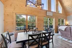 A wooden cabin interior with a dining table, chairs, a sofa, and large windows.