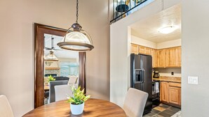 A dining area with a round wooden table, two chairs, and a potted plant. It leads to a kitchen with wooden cabinets and a black refrigerator. - Walk to Ski | Hot Tub | Great Location (Park City)