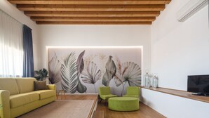 Een moderne woonkamer met een houten plafond, een groot bloemen behang, een gele bank, een groene fauteuil en een televisie op een houten plank.