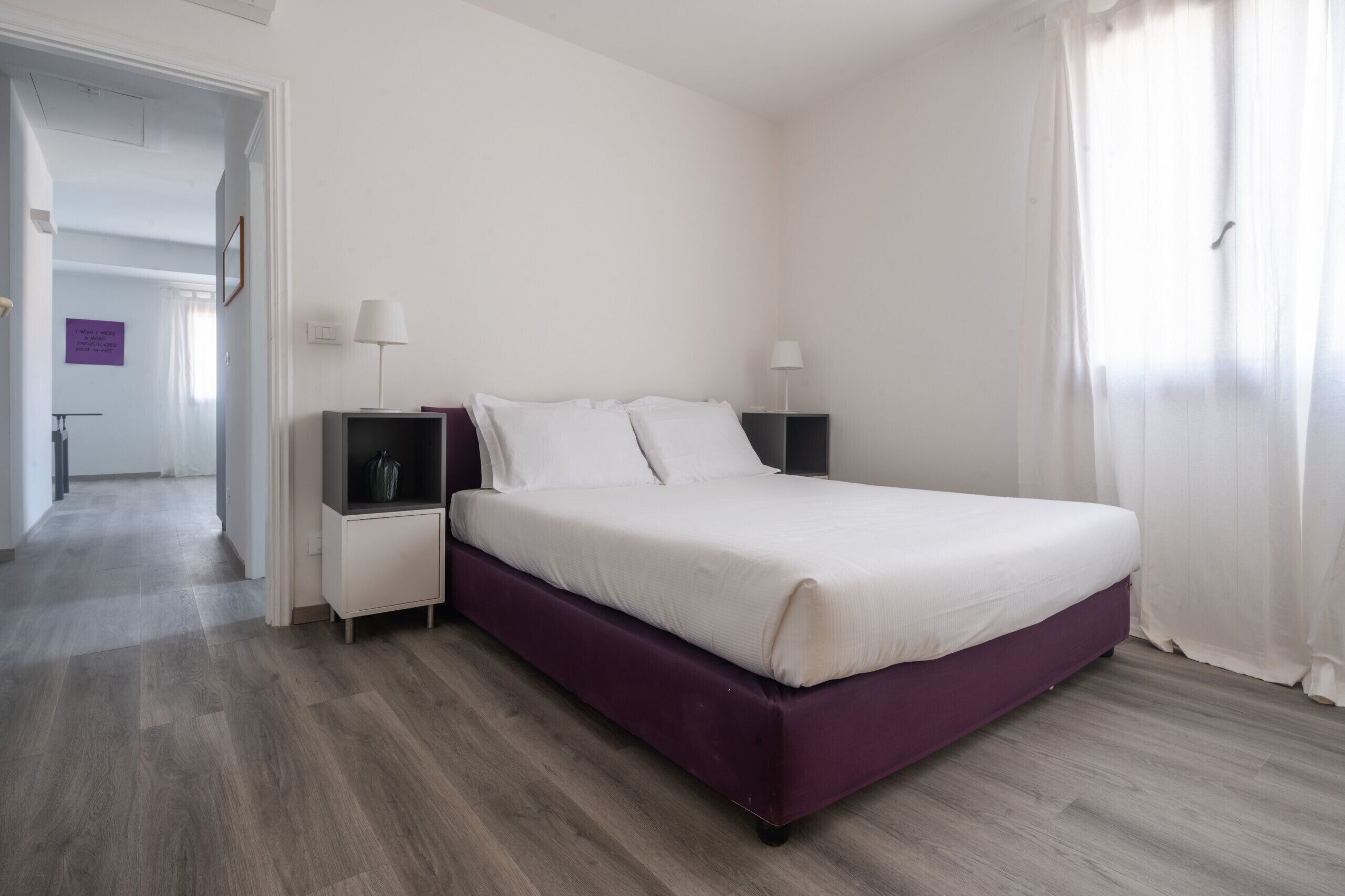 Une chambre à coucher avec un lit, deux tables de chevet, une commode, une lampe et une fenêtre avec des rideaux.