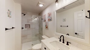 A modern bathroom with a glass shower enclosure, a white toilet, and a large mirror above the sink. - Fully Renovated Condo 786 Queens Grant (Hilton Head)
