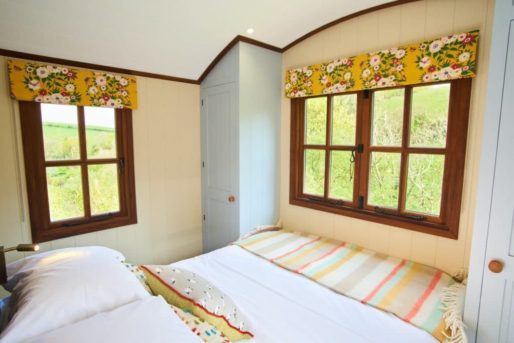 A bedroom with a bed, two windows with floral curtains, and a view of greenery outside.