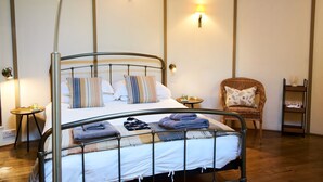 A bed with a metal frame, pillows, and folded towels. There are two bedside tables, one with a lamp and the other with a small table. A wicker chair is against the wall, and a shelf with decorative items is to the right. - Buzzard Treehouse -  a dittisham  that sleeps 5 guests  in 1 bedroom (Dittisham)