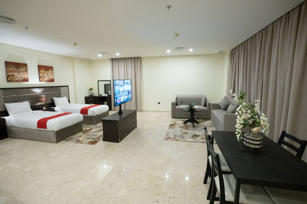 A hotel room with two beds, a TV, a sofa, a dining table, and a flower vase.