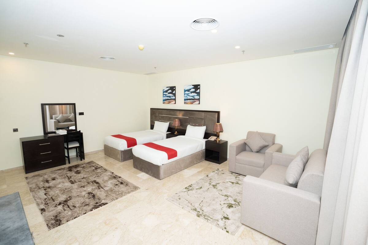 A modern hotel room with two beds, a desk, a chair, a sofa, and artwork on the walls.