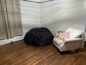 A corner room with a wooden floor, a black bean bag, a beige armchair with a patterned cushion, and a white wall.