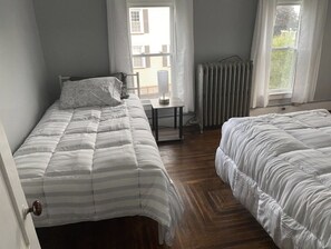 A bedroom with two beds, a radiator, a small table, and a window with curtains.
