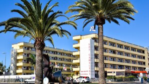 A multi-story hotel with a yellow facade, palm trees, and a large sculpture in front.